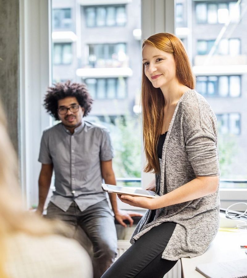 Afro-haired man and auburn-haired woman holding a tablet device chat to a colleague as they perch on desks by a window.